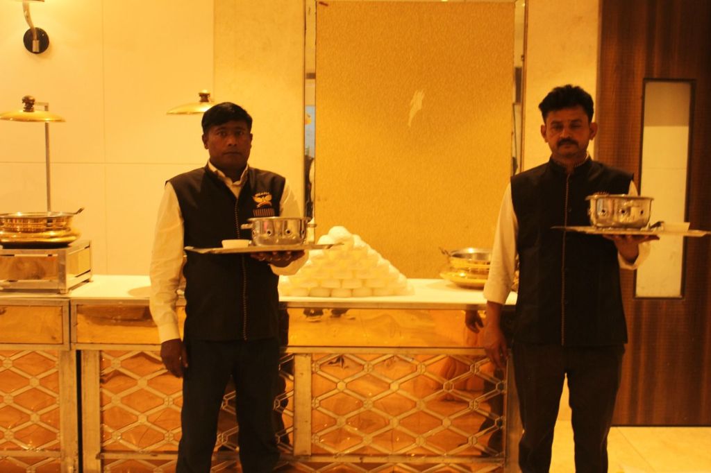 Two Mithila Events waiters in uniform holding serving trays at a buffet counter