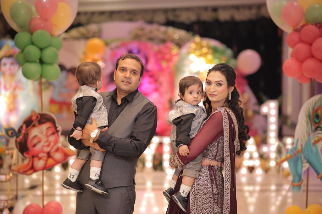 Parents holding their twins in front of the Krishna theme first birthday stage in Noida